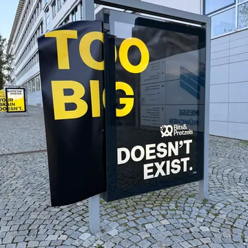 Black-and-yellow Bits & Pretzels sign reading “Too big doesn’t exist” in front of a building