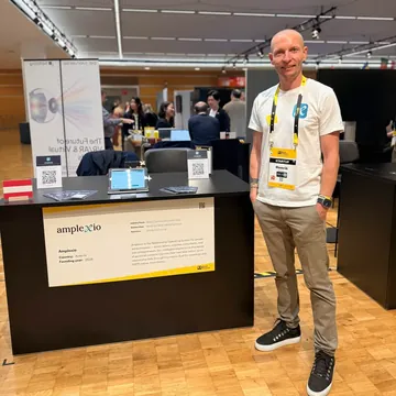 Henrik Gruber in a white Amplexio T-shirt in front of booth 0–62 in the exhibition hall
